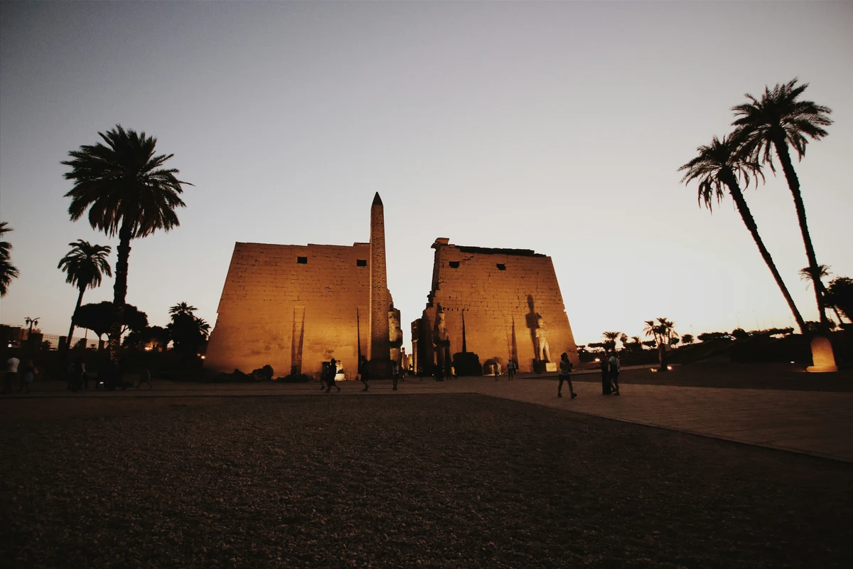 The illuminated pylon and obelisk of Luxor Temple at dusk with palm trees.
