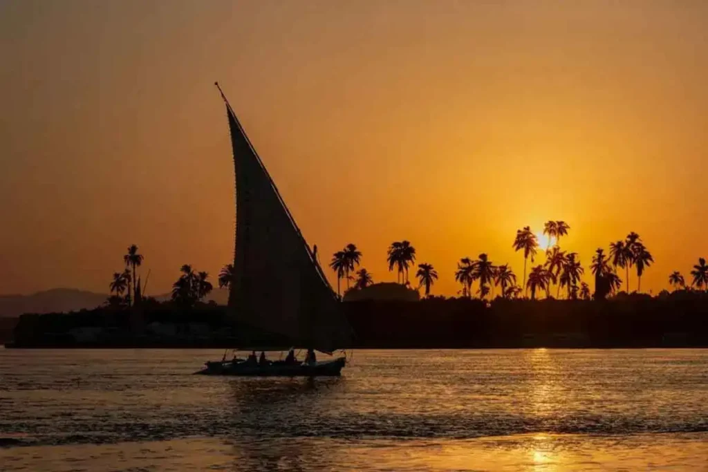 A sailboat, a felucca, on the Nile River at sunset, with palm trees appearing as shadows against the golden sky.
